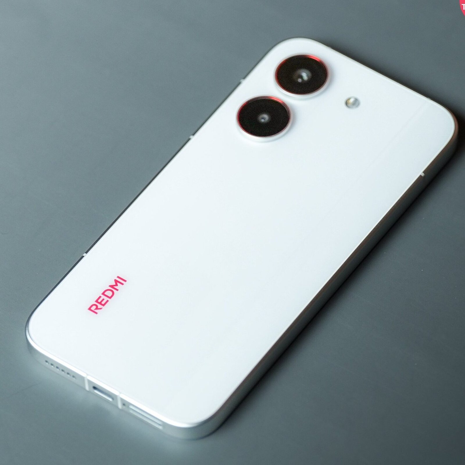 Redmi Turbo 5 Specs and Launch Date Leak 12 White Redmi smartphone with dual rear cameras and minimalist design placed on a dark grey surface.